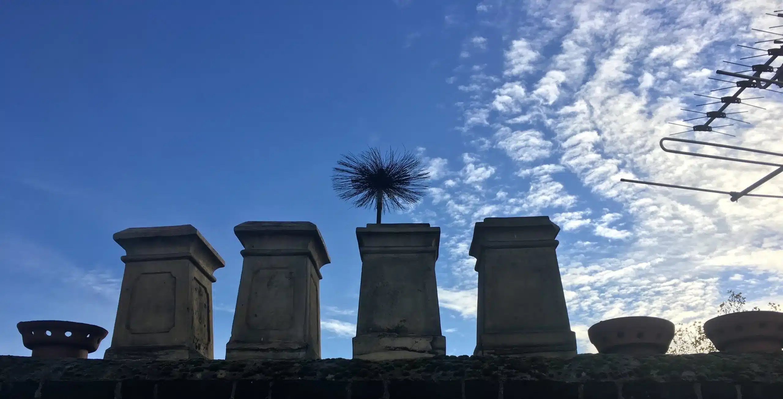 chimney-brush-chimney-pots-sweepsmart Chimney sweep brush extending above a row of chimney pots against a blue sky